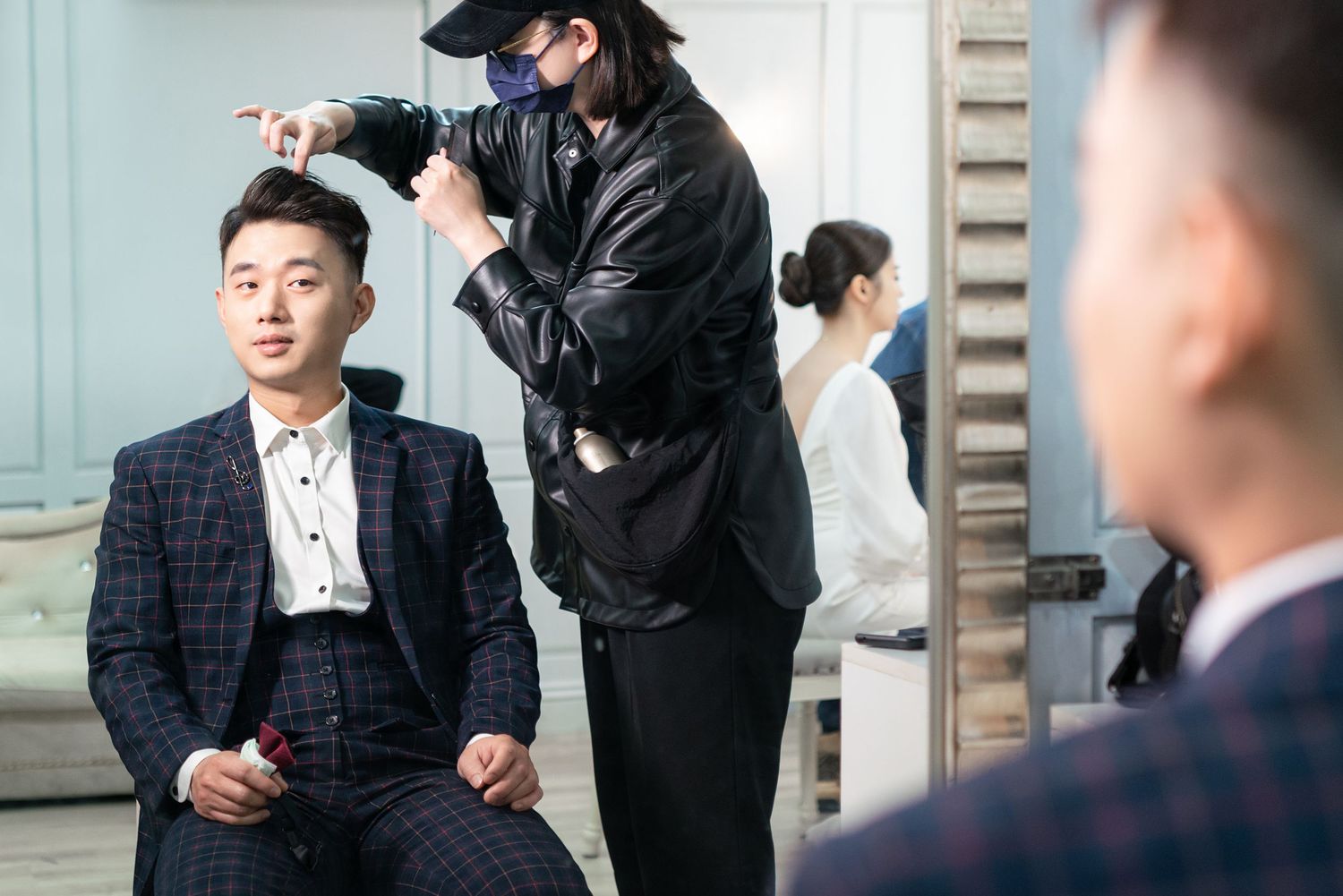 A stylist wearing black and a face mask works on fixing a client's hair as they sit in a plaid suit.