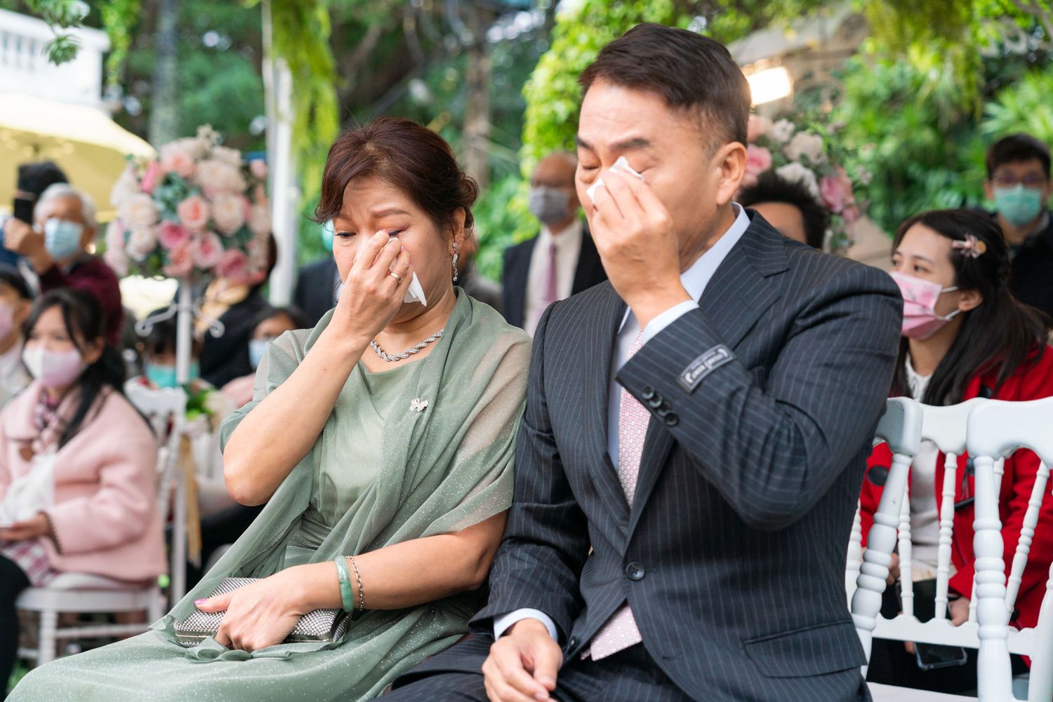 People attending an outdoor event react emotionally by covering their mouths while seated in white chairs under greenery.