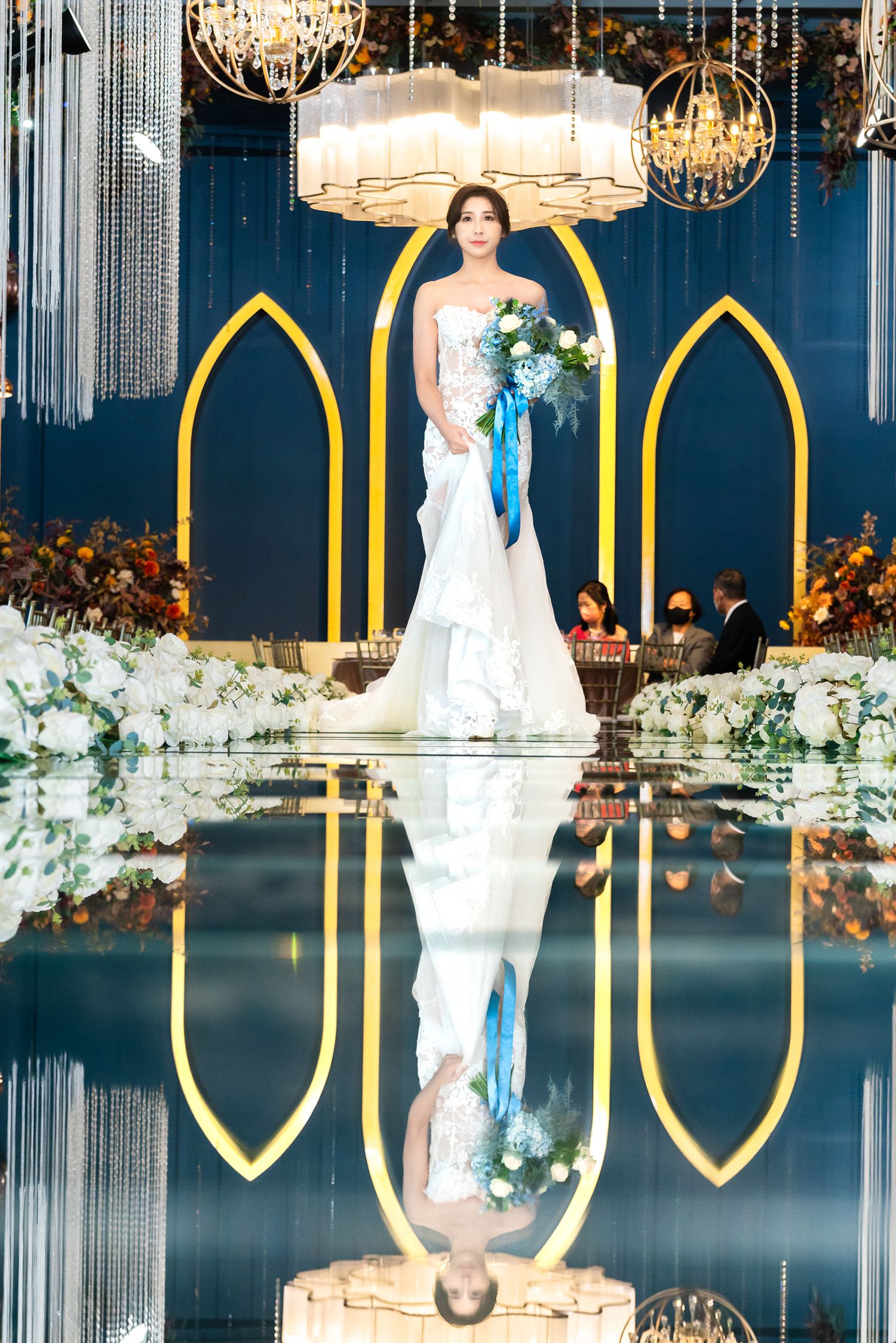 A bride in a white mermaid gown holds a blue-ribboned bouquet under elegant chandeliers with gold arches reflecting below.