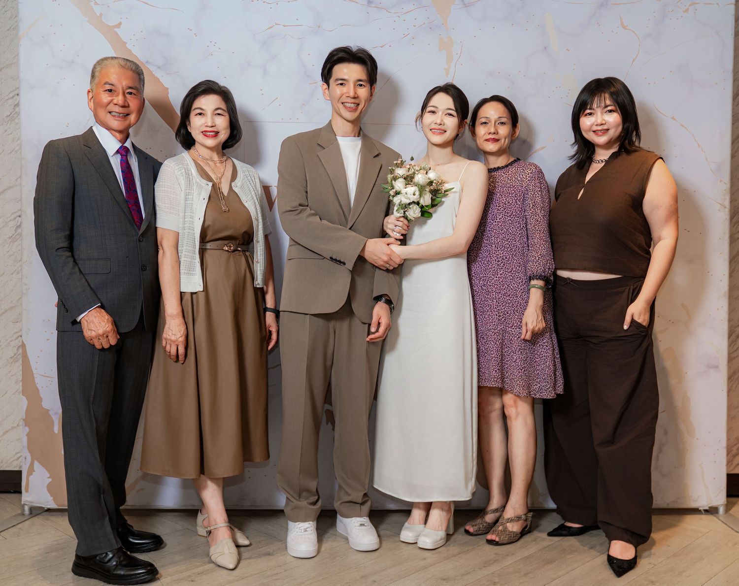 A group of six people in formal attire pose together for a wedding photo against a neutral backdrop.