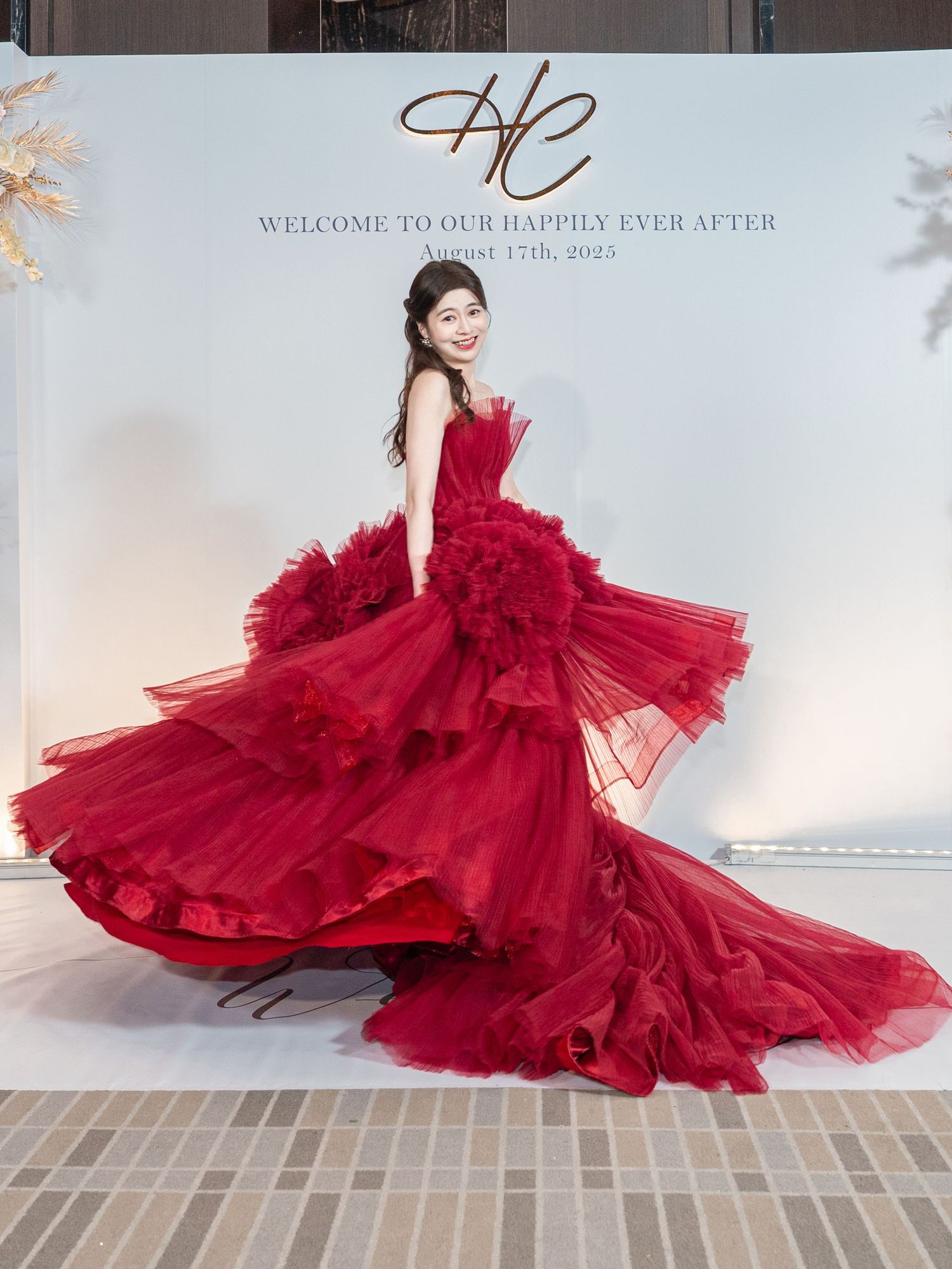 A person in a stunning red tulle ballgown twirls in front of an elegant wedding backdrop with script lettering.
