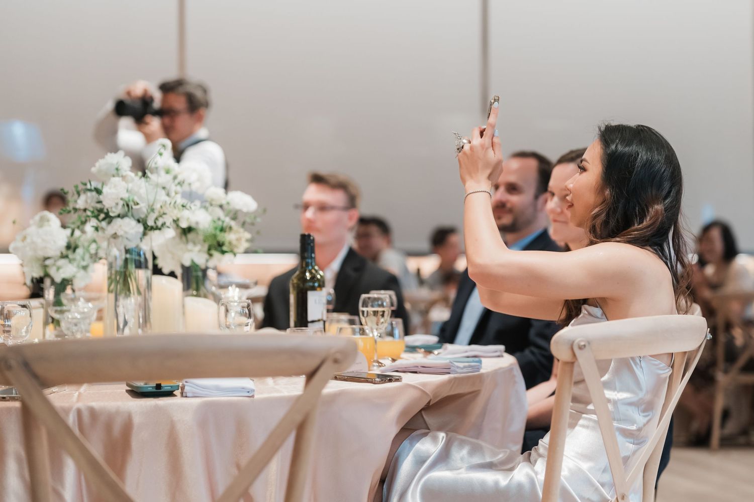 People dining at elegant wedding reception with white tablecloths and floral centerpieces in a formal venue.