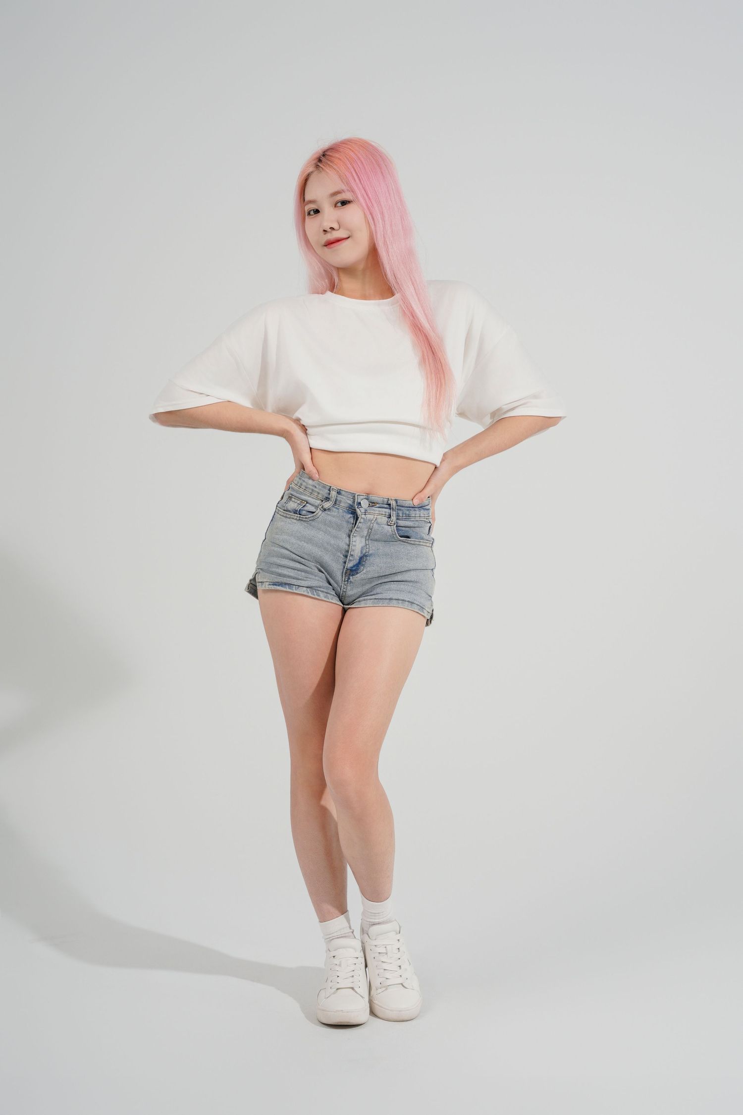 Person in white crop top and denim shorts with pink hair poses against white background in casual summer outfit.