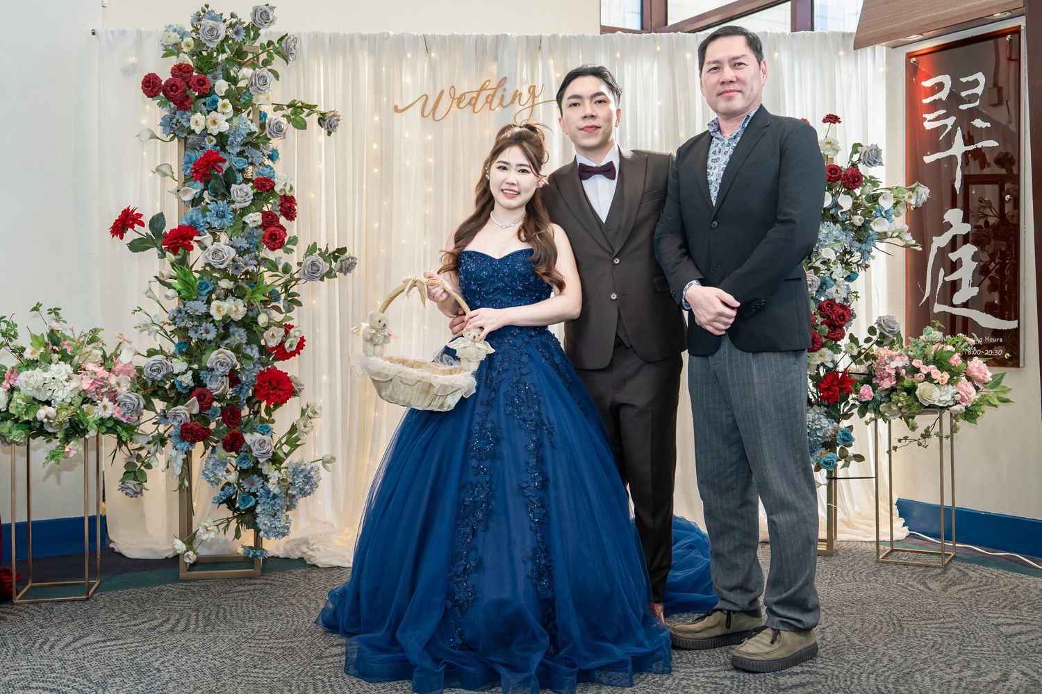 A couple poses in front of a rustic wall with blue roses, wearing elegant blue and gray formal attire.