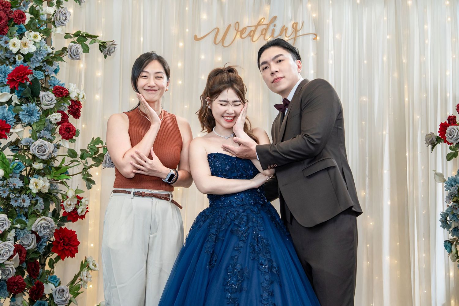 Couple in formal attire dancing and posing playfully in front of white backdrop with red floral decorations.