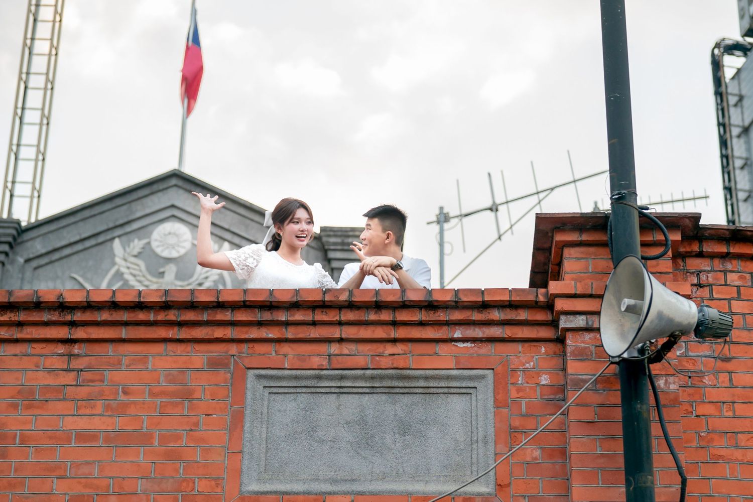 Two people on a ladder installing a security camera on a brick wall exterior.