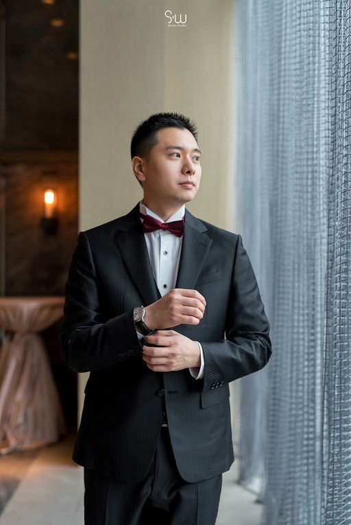 A formal wedding portrait shows a groom in a black tuxedo and bow tie adjusting his cuff while gazing out a window.