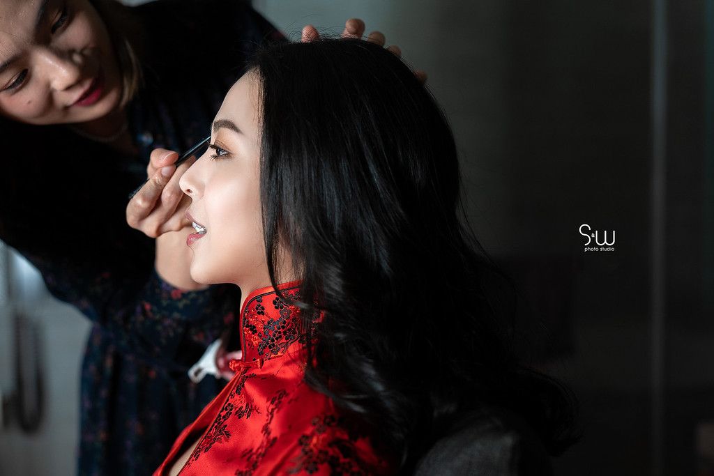 Makeup artist applies cosmetics to a bride wearing a red traditional Chinese dress.