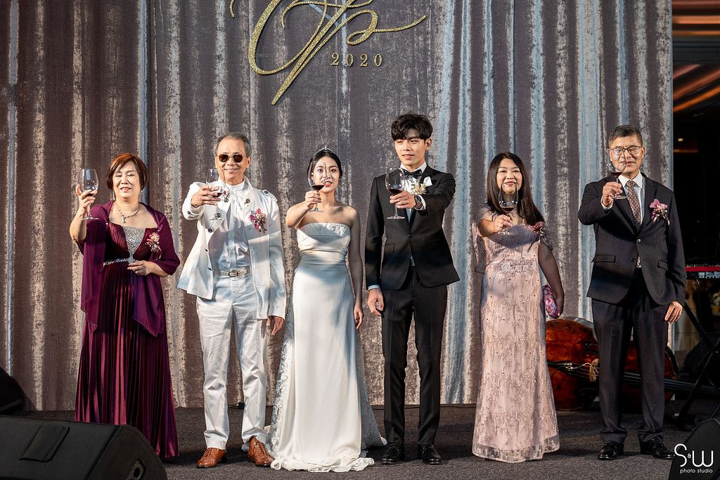 Wedding party group poses together in formal attire against silver backdrop.