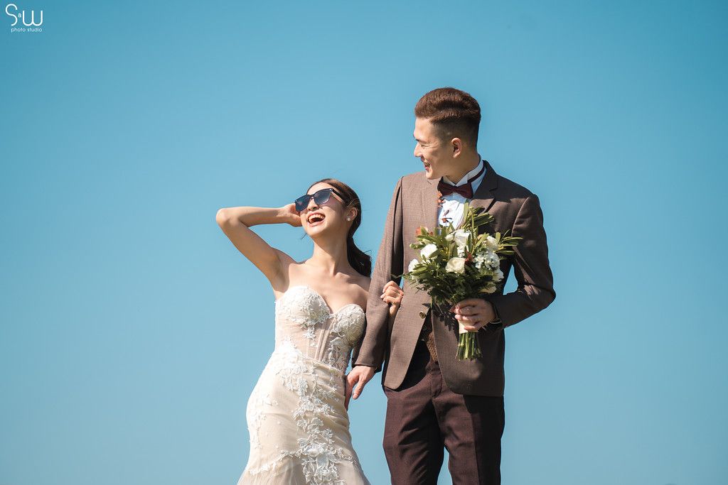 Joyful wedding portrait against bright blue sky with white bouquet.