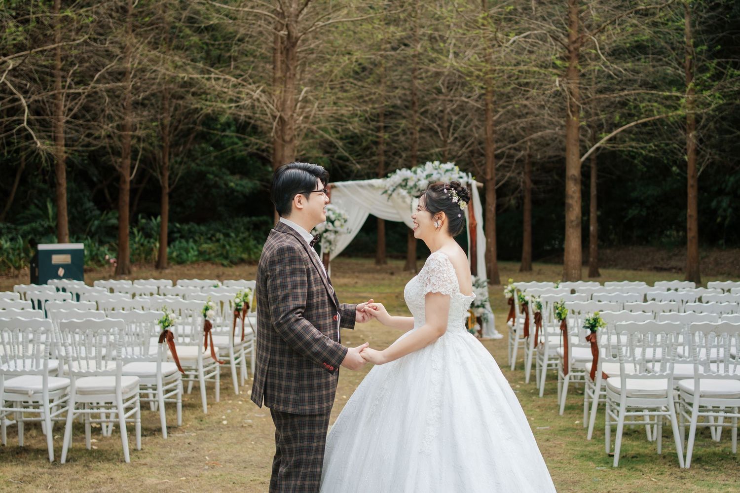 An outdoor wedding ceremony setup in a wooded area with rows of white chairs and a couple standing at the altar.