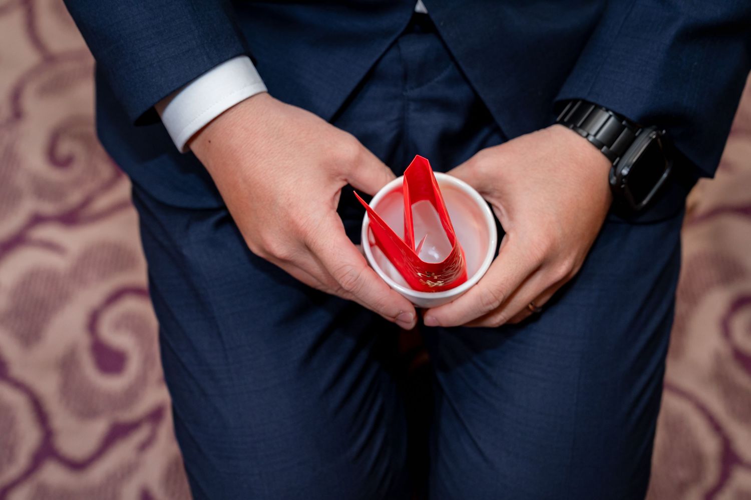 Groom in a navy suit and black wristwatch holds a small white cup with red envelopes during a wedding tea ceremony.