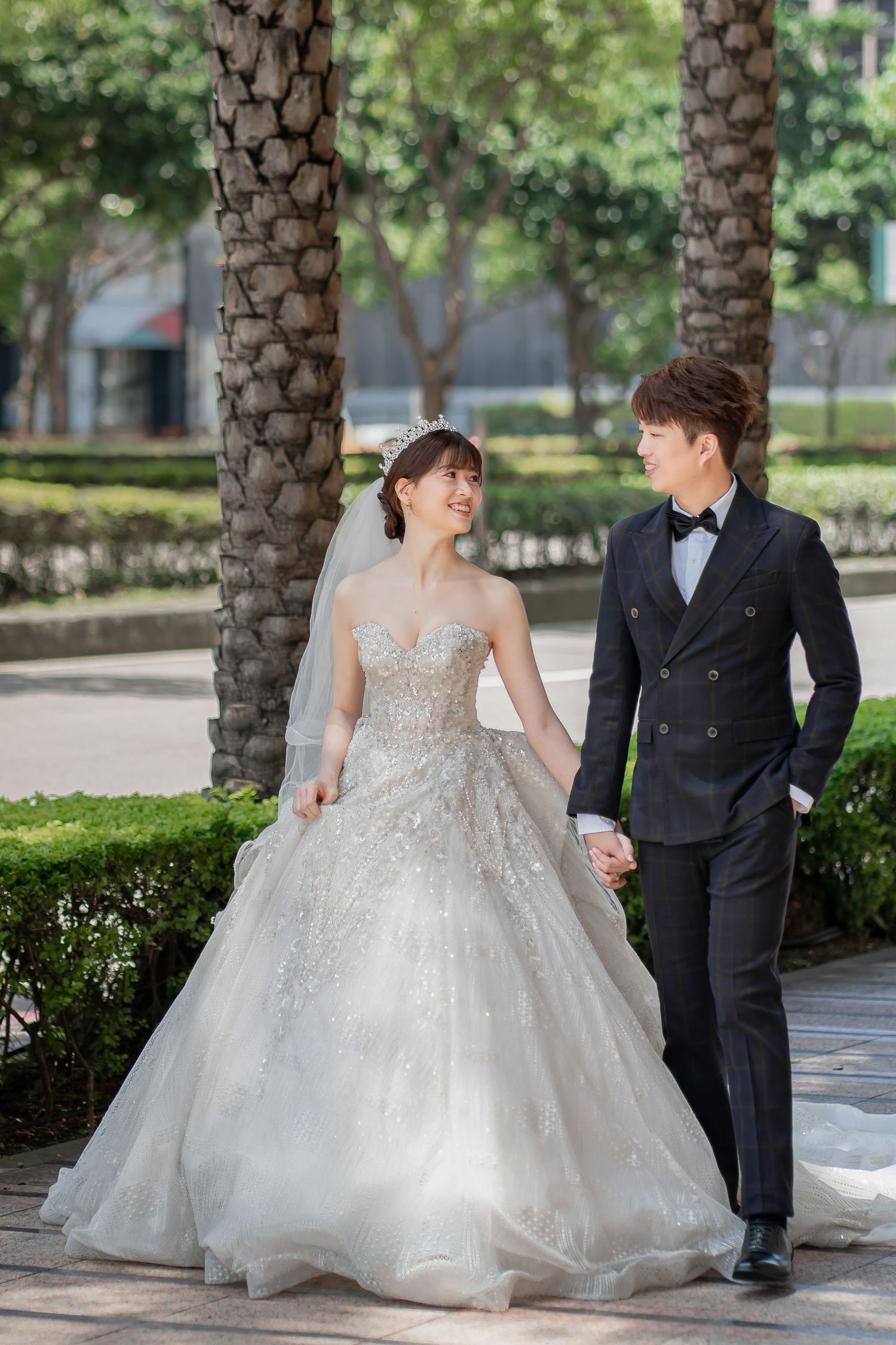 A couple in wedding attire walks hand-in-hand outdoors, bride in a beaded gown and groom in a black tuxedo.