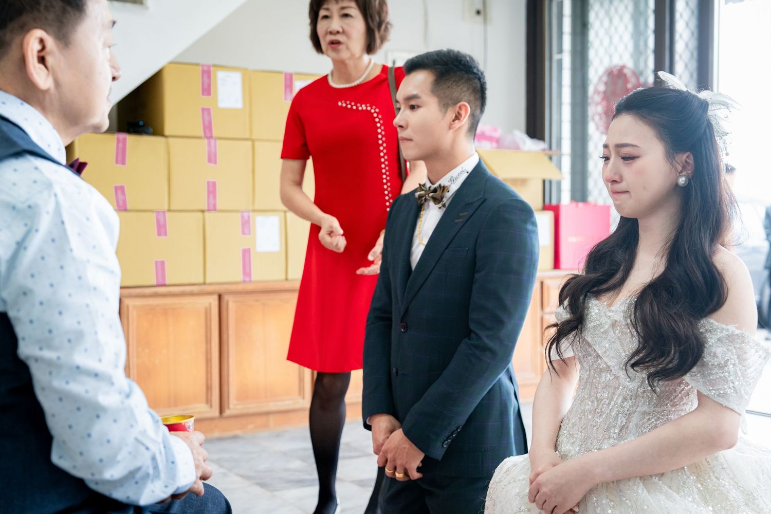 A couple in wedding attire stands with guests indoors, the bride in a white gown and groom in a suit with bow tie.