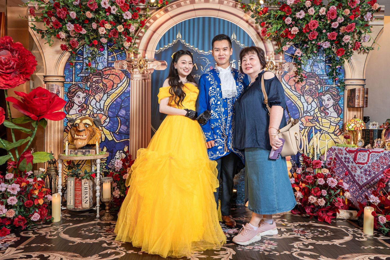 Three people posing together in a Beauty and the Beast themed setting with floral decor and stained glass backdrop.