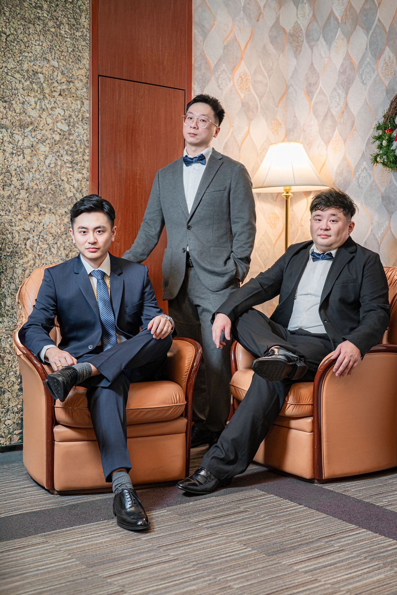 Group of men in formal suits and bowties seated on brown leather chairs in an elegant hotel setting.