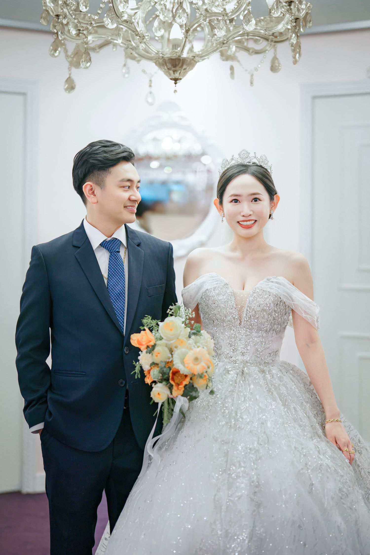 Elegant wedding couple poses under crystal chandeliers in formal attire with peach and white floral bouquet.