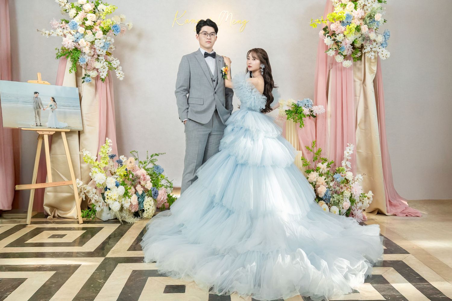 A romantic wedding photoshoot featuring a couple in a blue gown and grey suit against a pink floral backdrop.