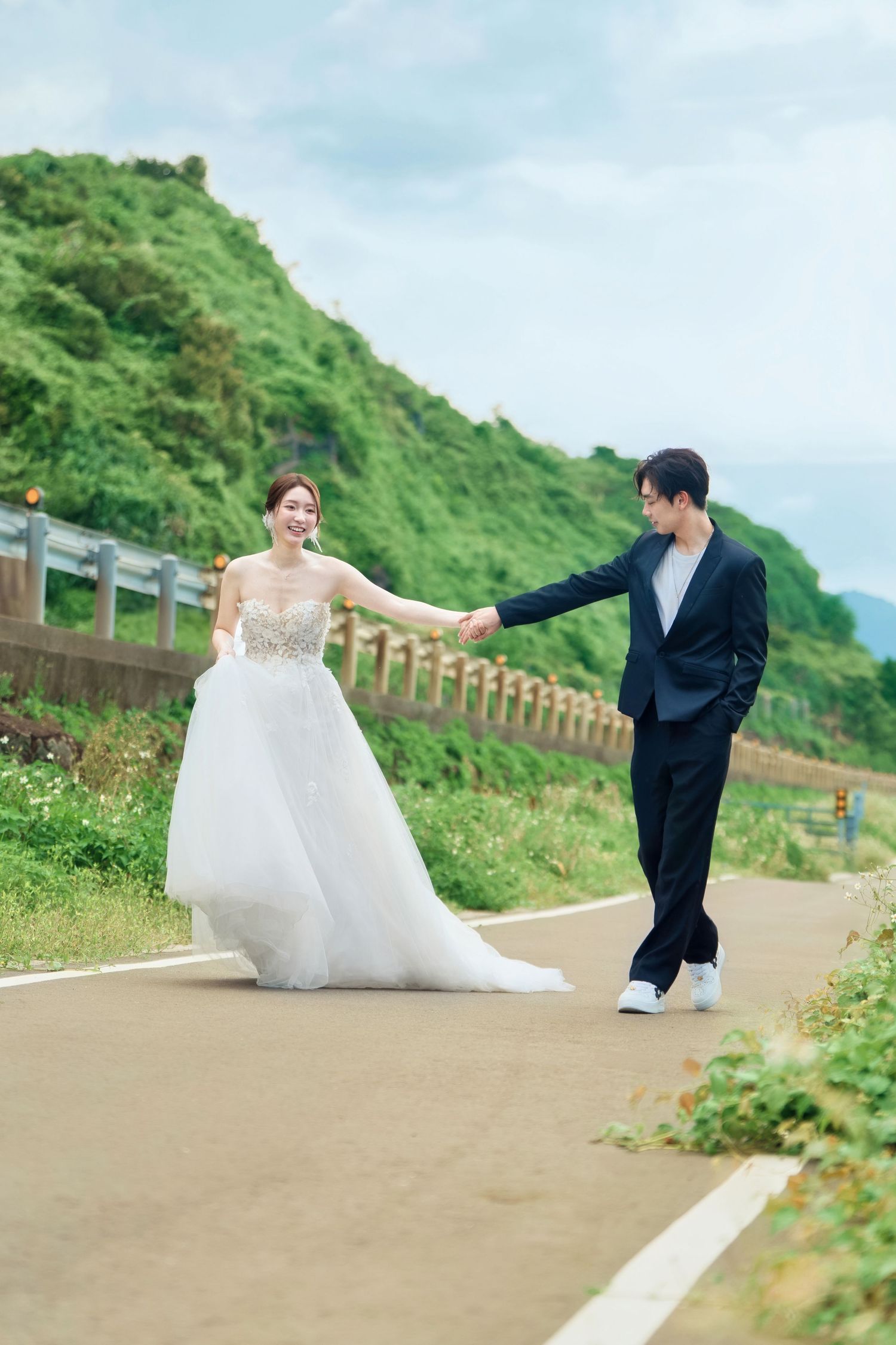 A couple in wedding attire playfully poses for photos on a coastal path with lush green hills in the background.