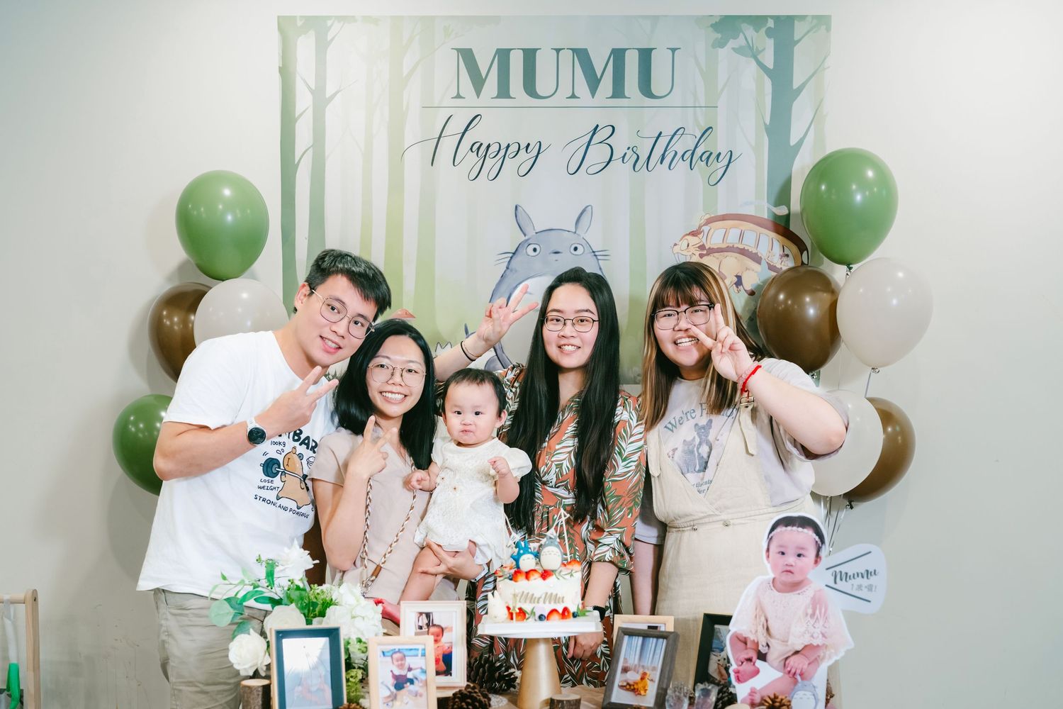 Birthday celebration with green and white balloons, woodland forest theme decorations, and party guests posing with cake and gifts.