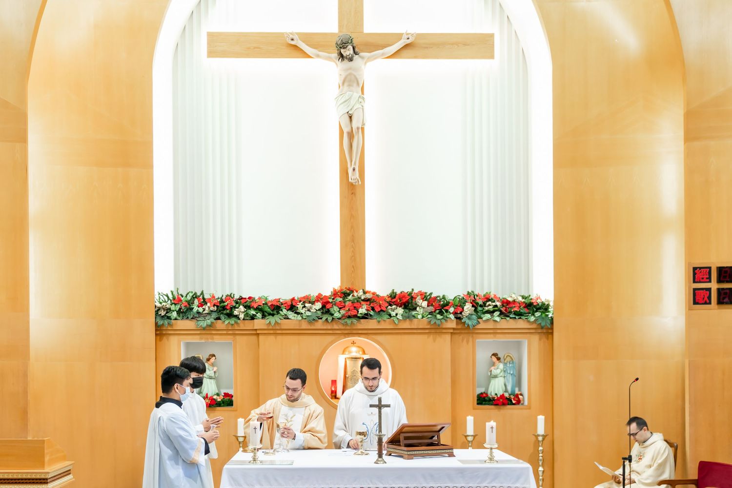 Priests conducting a ceremony at an altar decorated with red and white floral arrangements.