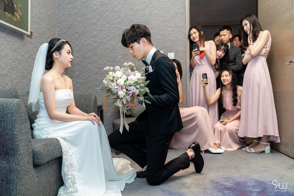 Groom kneels before bride while bridesmaids in pink dresses watch from behind.