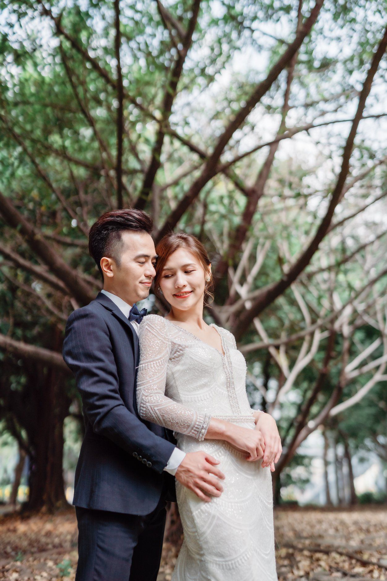 Romantic couple embrace in formal wear under branching trees.