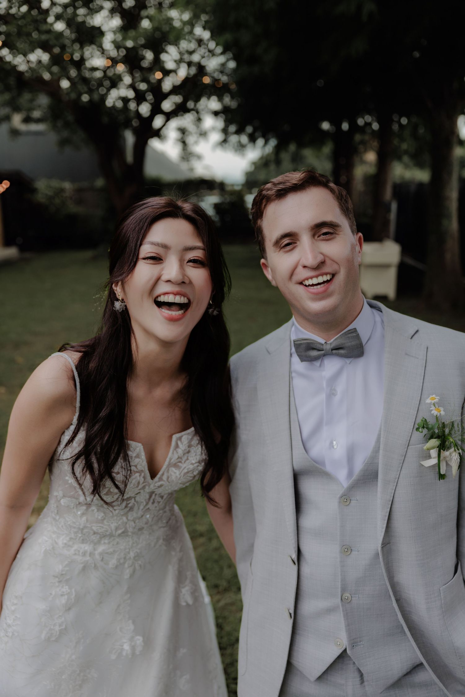 Joyful bride in white lace gown and groom in light gray suit with bow tie laughing together outdoors at wedding reception.