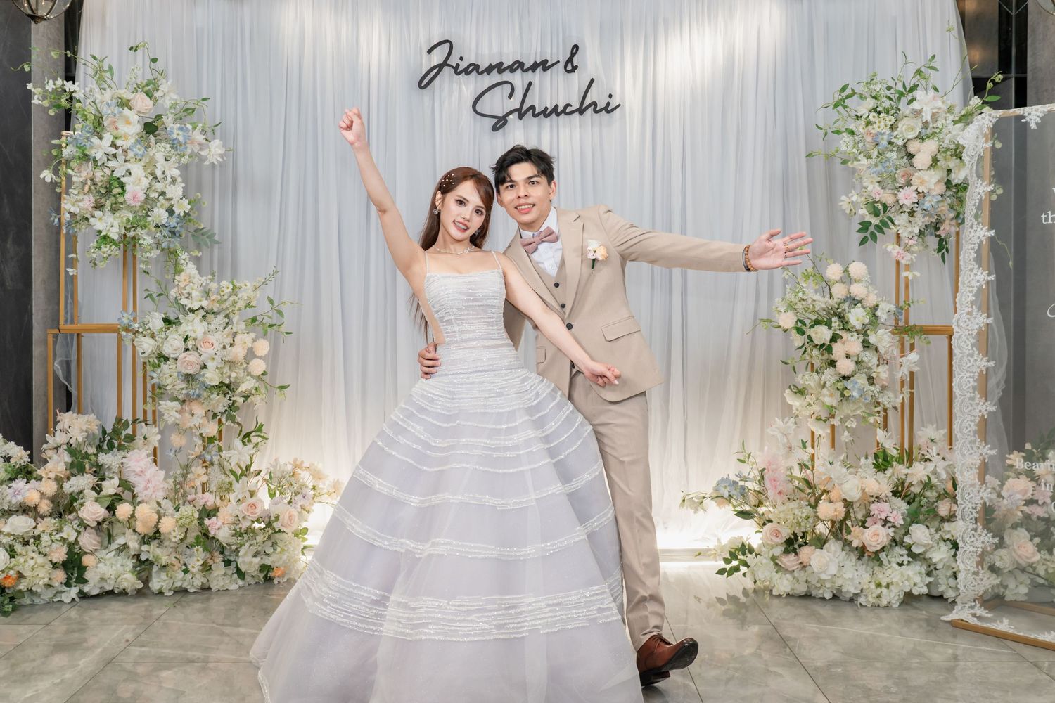 A joyful couple poses in wedding attire before a floral backdrop with their names Jianan and Shuchi displayed.