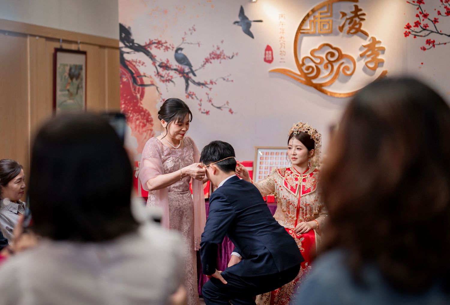 Groom kneeling before a bride in a red embroidered gown during a traditional Chinese wedding tea ceremony as a woman and guests look on.