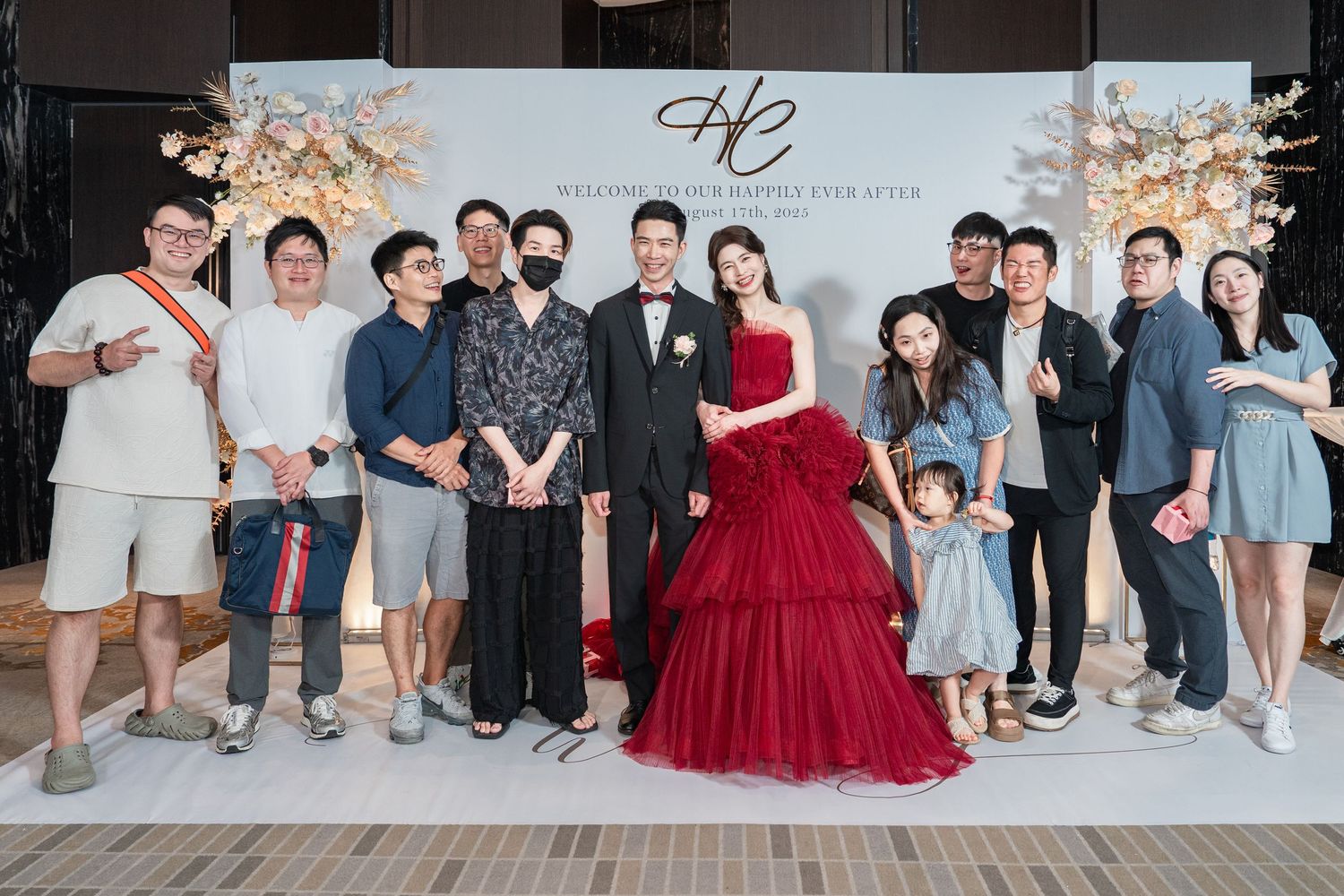 A celebratory group photo at a wedding reception with a couple in black tuxedo and red gown in front of a decorative backdrop.