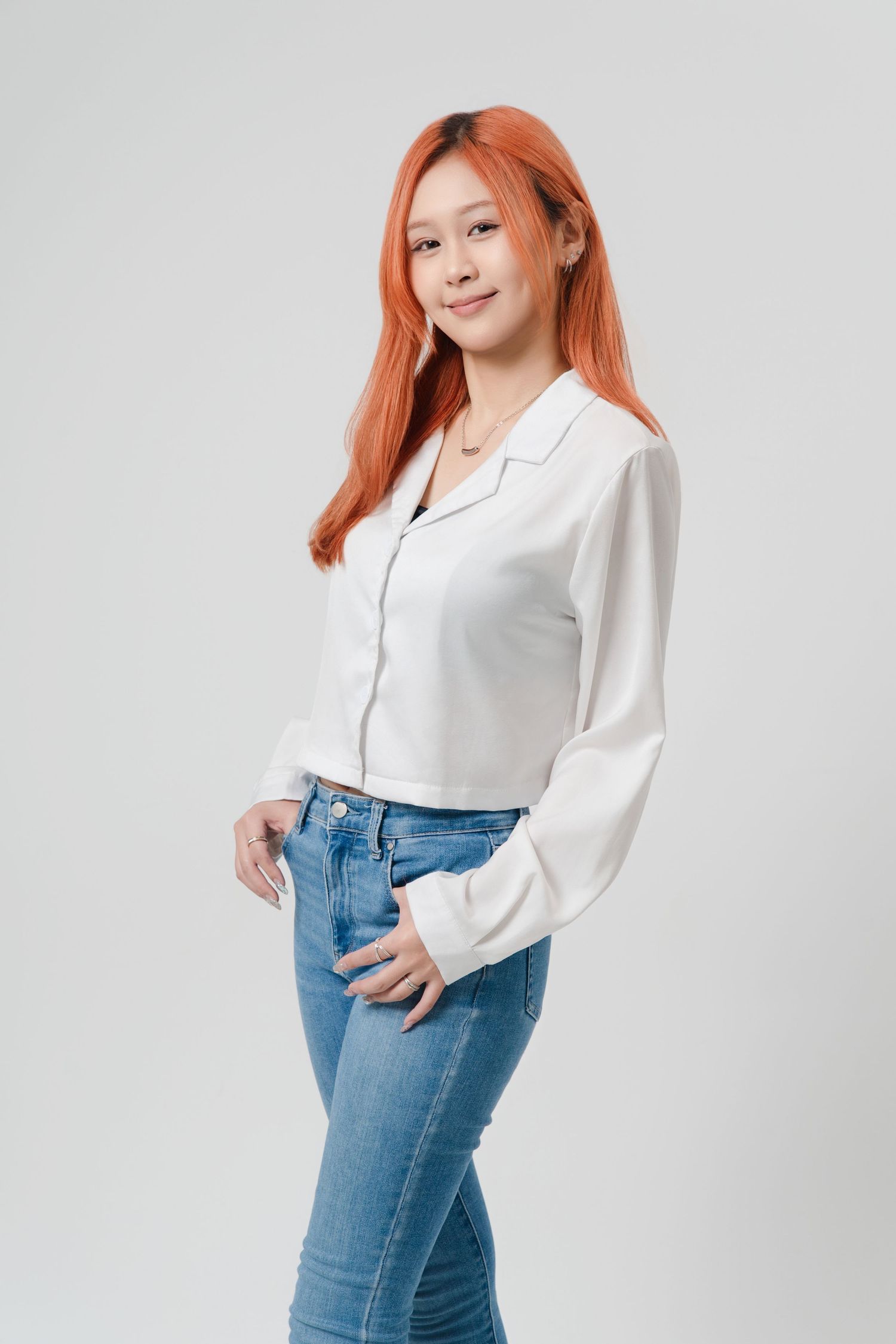 Young woman with red hair wearing white button-down shirt and blue jeans poses in studio against light background.