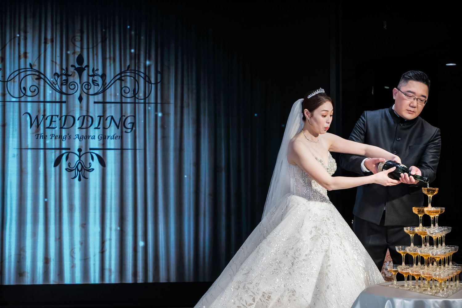 Wedding couple pours champagne into elegant tower of glasses at luxurious reception venue.