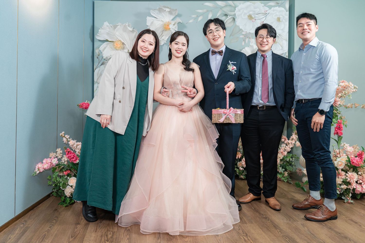 A group photo at a wedding celebration with pink floral decorations against a teal wall.
