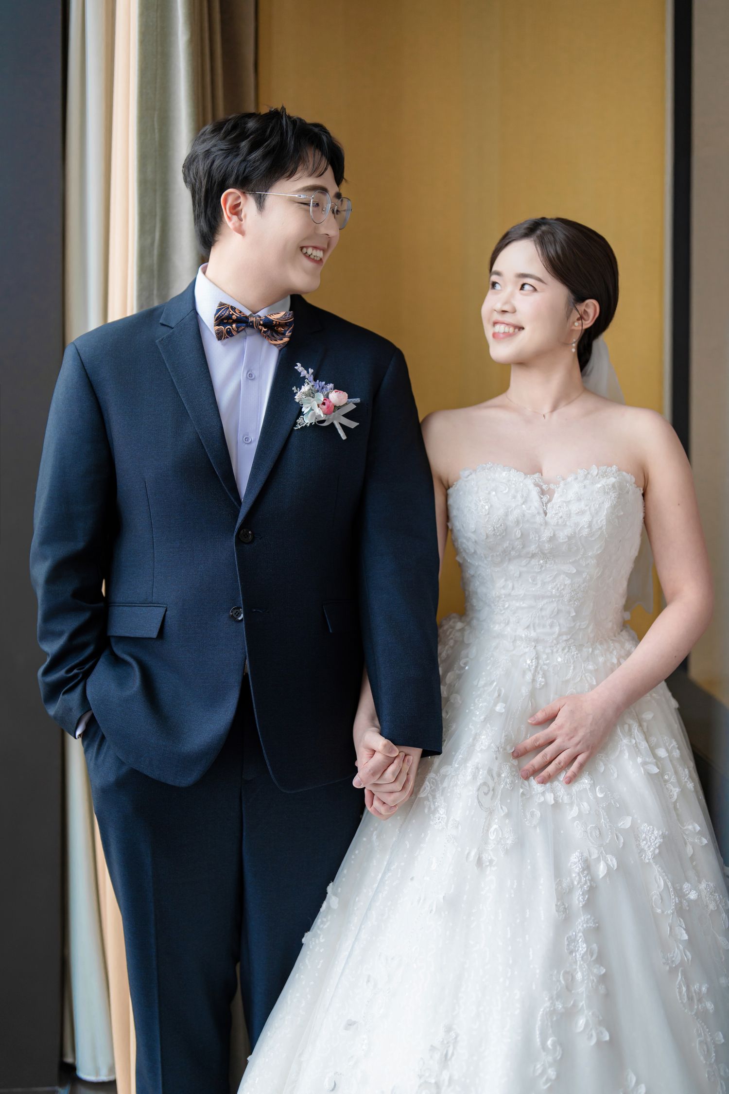 A newlywed couple shares a candid moment together in formal wedding attire against a neutral background.