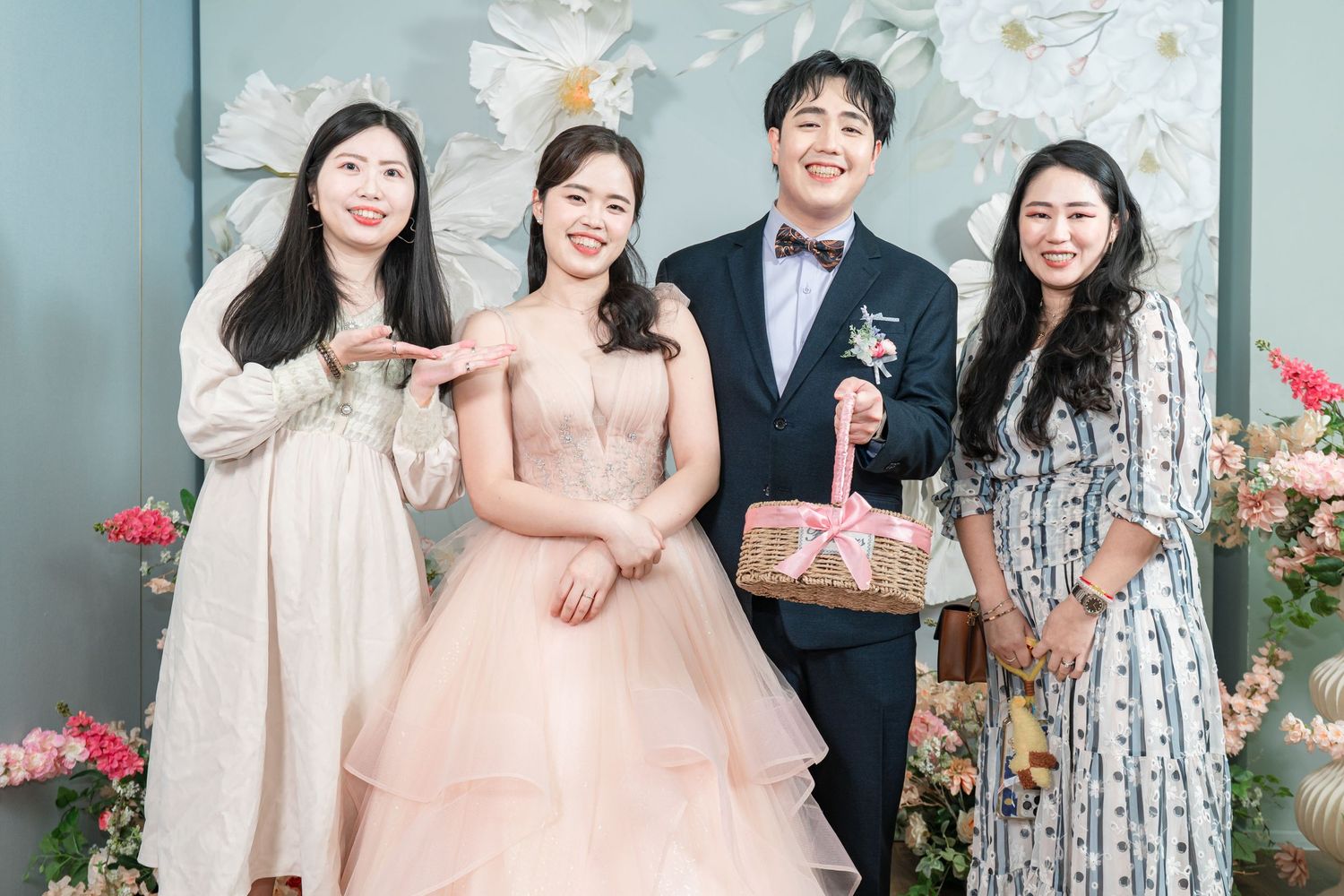 A group of people in formal attire pose together at a wedding celebration with floral decorations in the background.