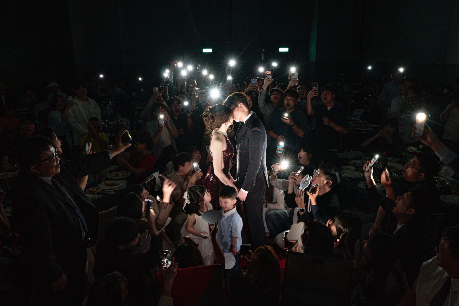 Couple shares romantic moment surrounded by crowd holding phone lights at a dark evening celebration.