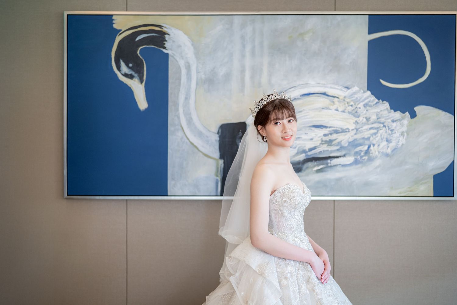 Bride in strapless white gown and tiara poses elegantly before a blue swan painting backdrop.