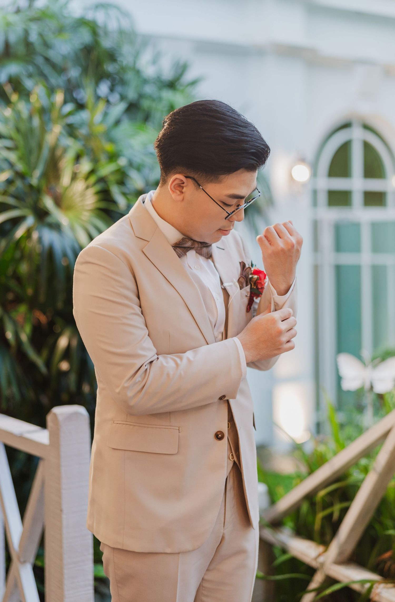 A groom in a beige suit and bow tie adjusts his boutonniere against a backdrop of greenery and arched windows.