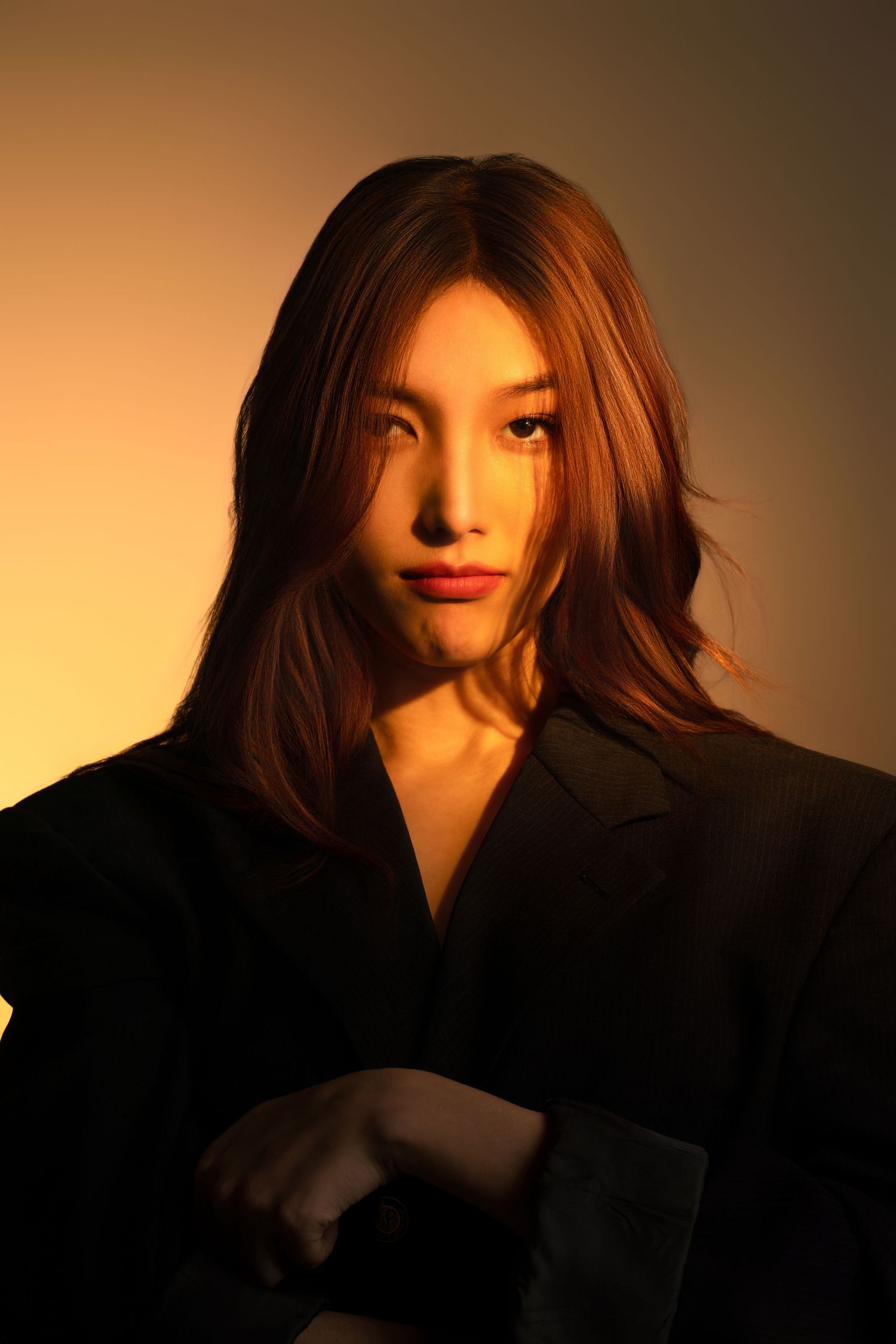 A dramatic portrait lit by warm amber lighting shows a person in a black blazer with flowing brown hair against a gradient background.