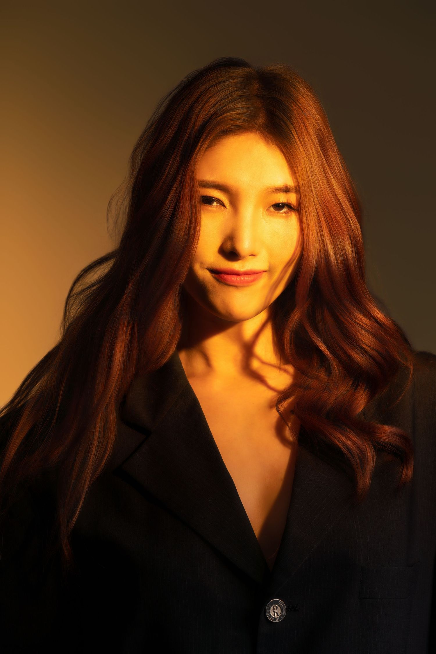 Portrait photo in warm lighting showing a person with long wavy brown hair wearing a black blazer against golden background.
