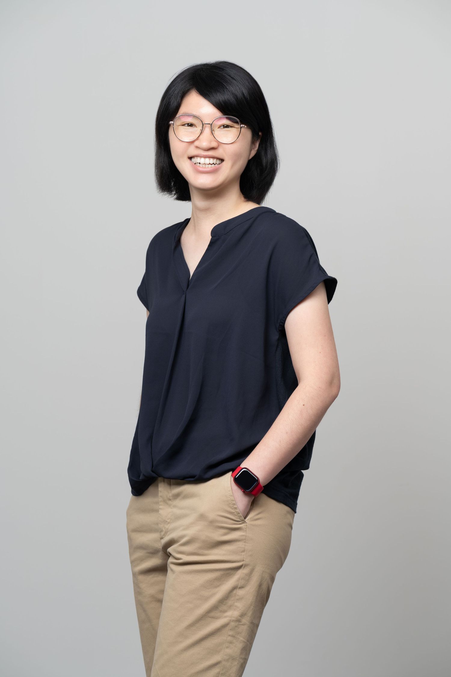 Professional headshot of someone wearing glasses and a navy top with khaki pants against a gray background, smiling warmly.
