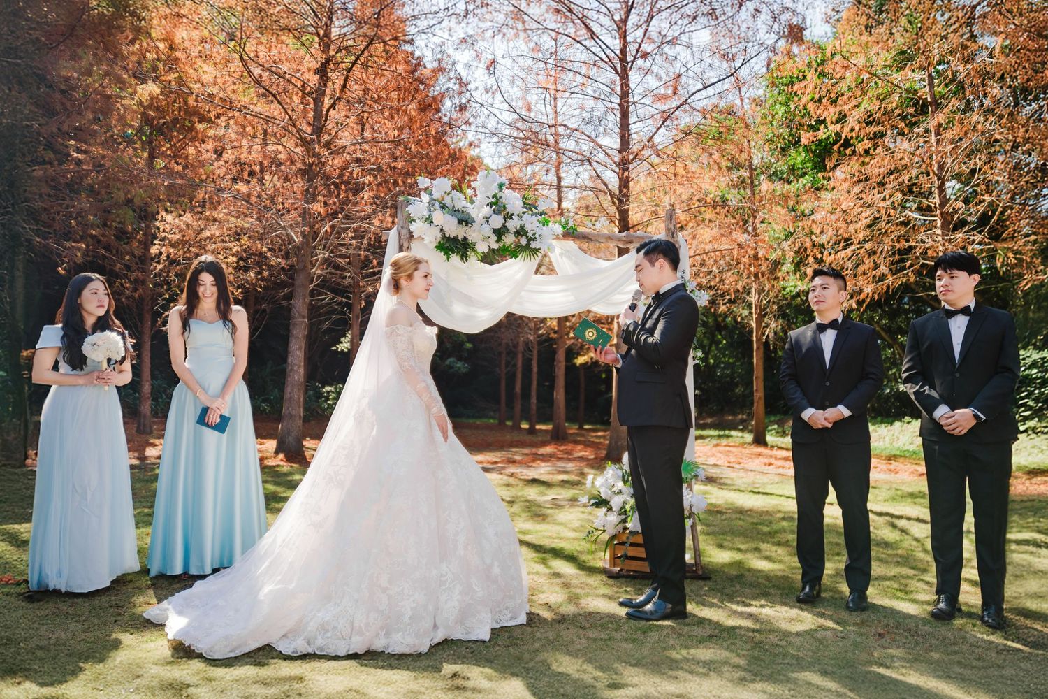 A wedding ceremony takes place outdoors in autumn with bridesmaids in blue dresses and groomsmen in formal suits.