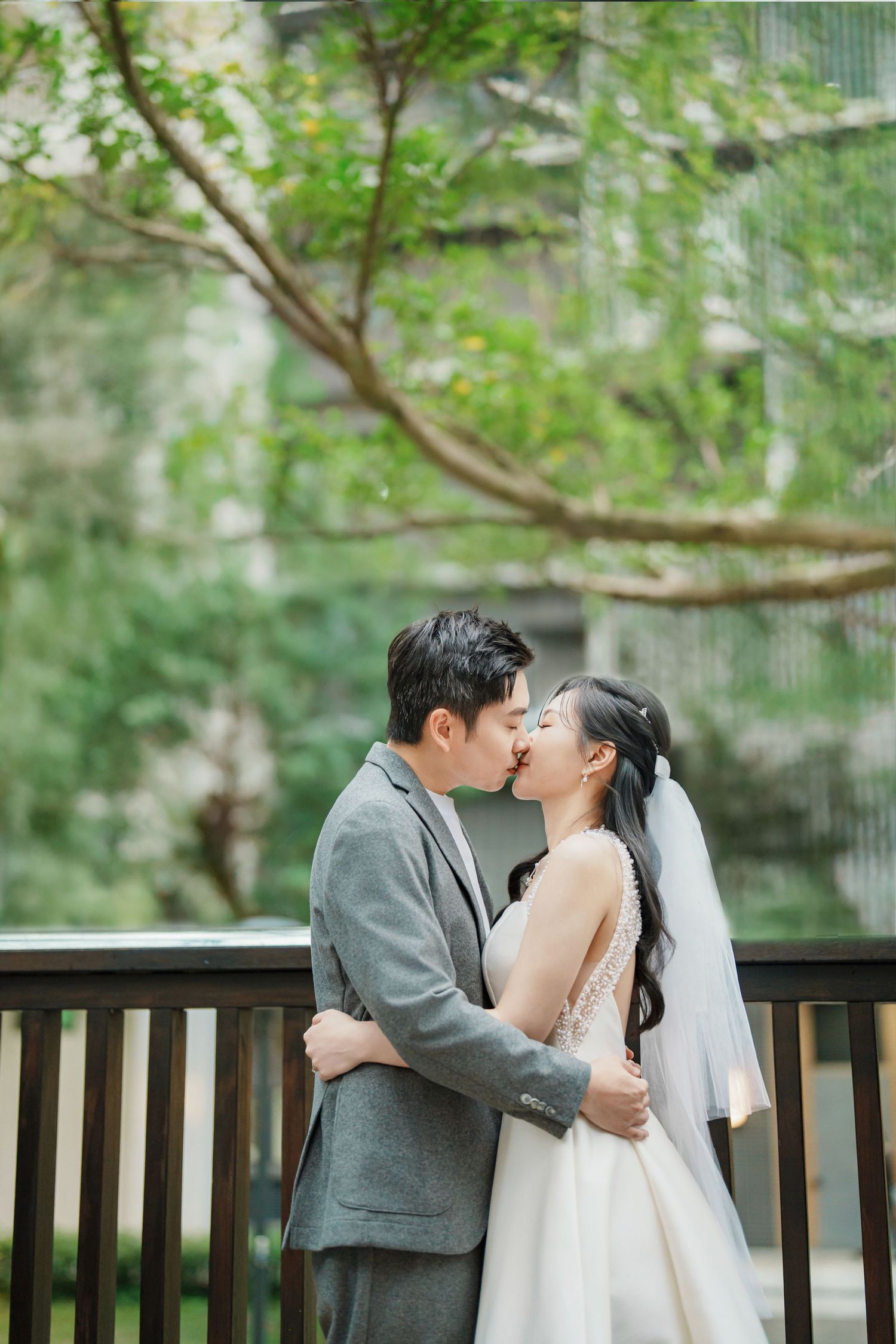 A romantic wedding couple shares tender kisses on a wooden bench surrounded by lush green foliage.