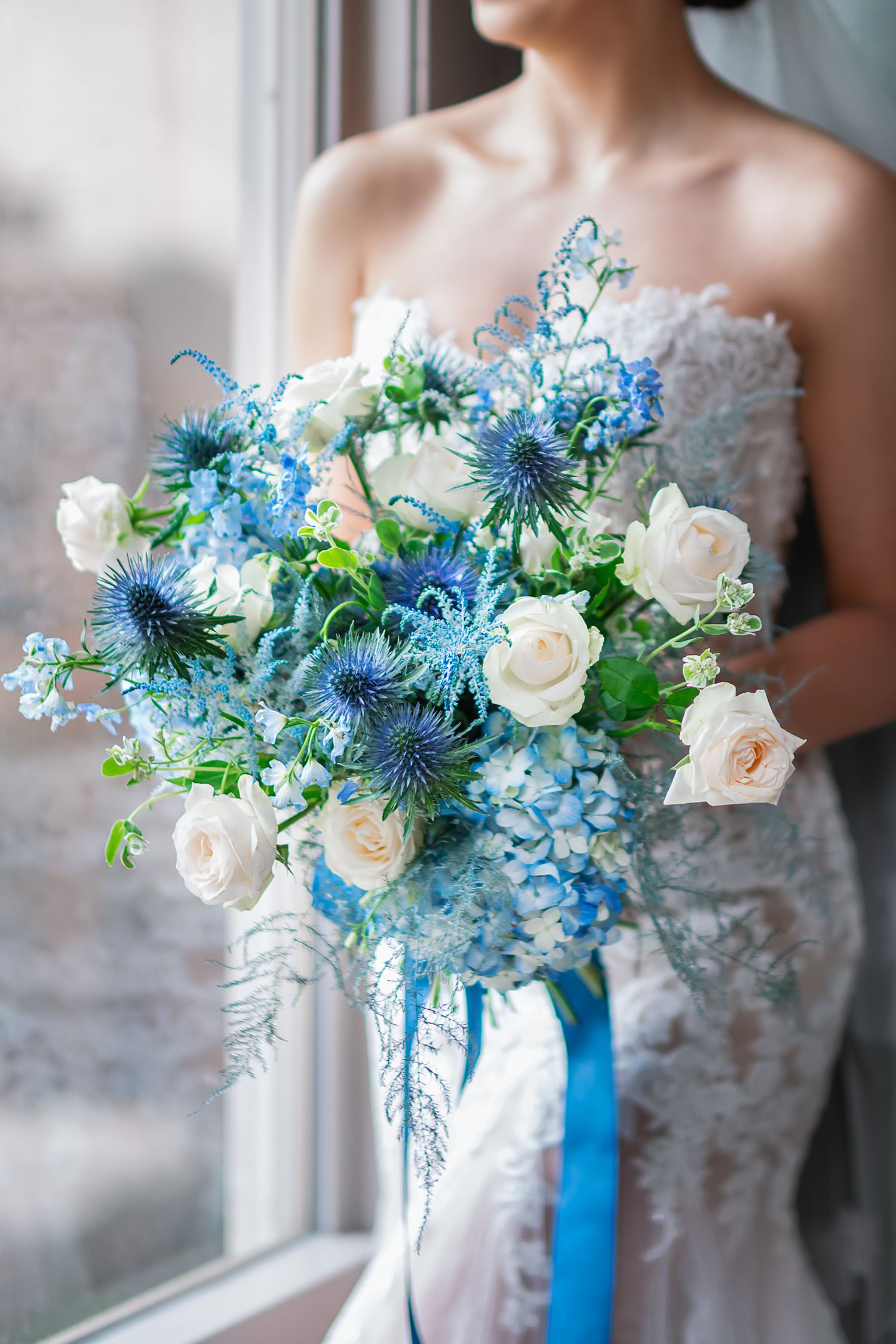 A stunning bridal bouquet featuring white roses, blue thistle flowers and delphinium with a blue ribbon wrap.