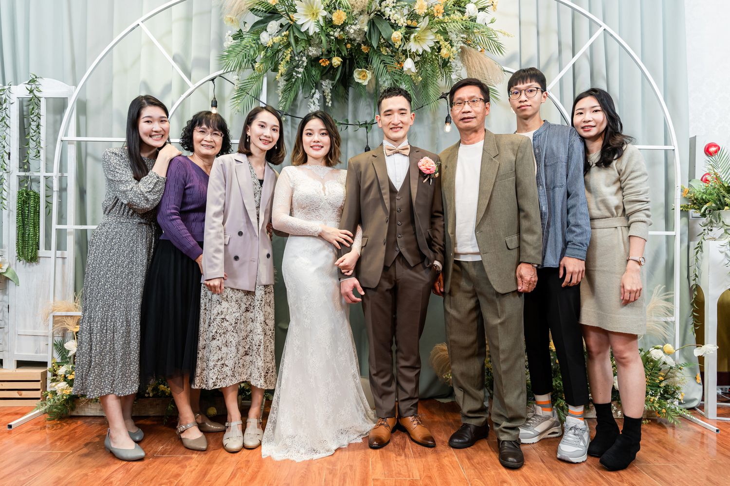 Wedding party poses together under floral arch at indoor reception venue.