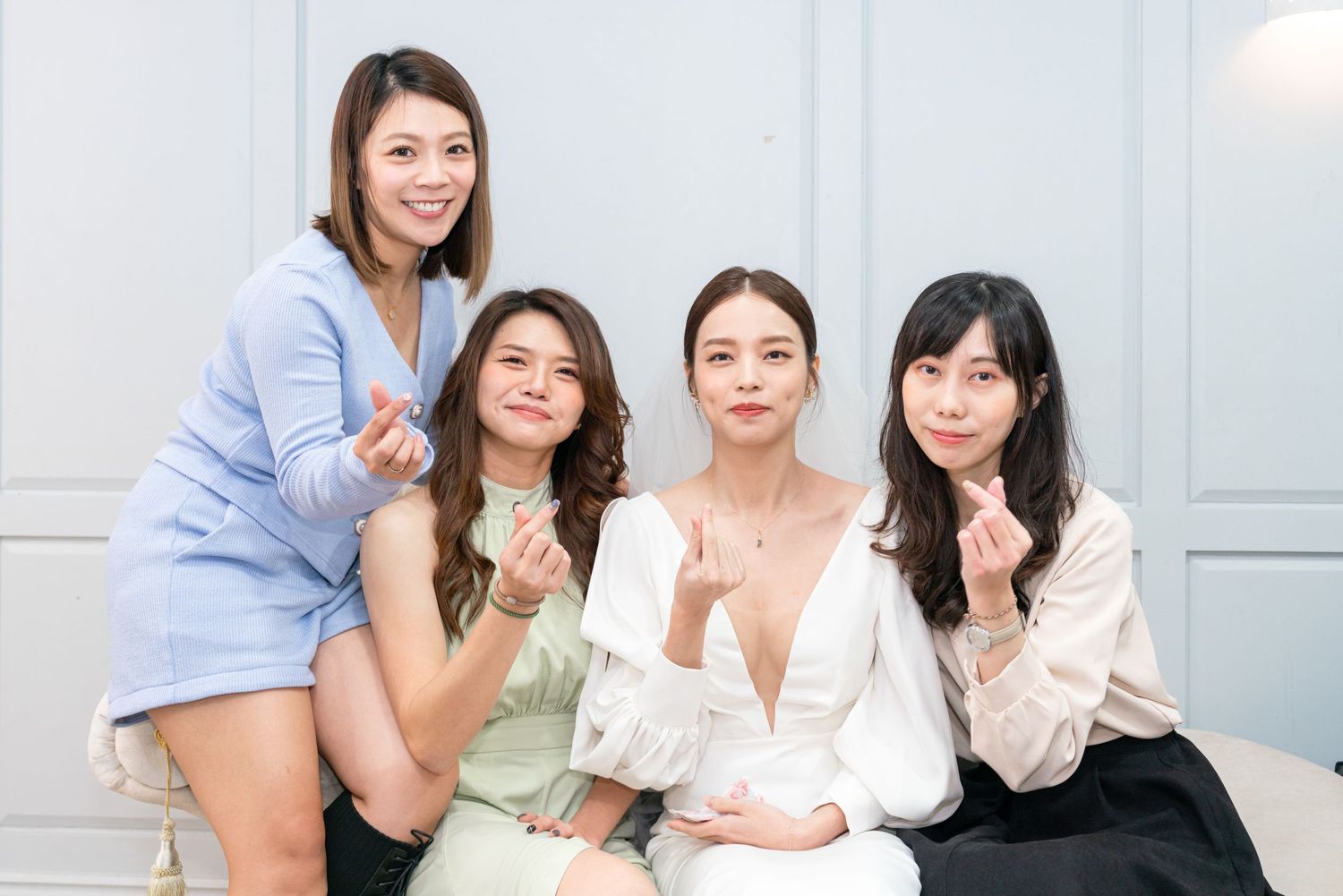 Four friends sitting together on a sofa while wearing light colored casual outfits and making heart gestures.