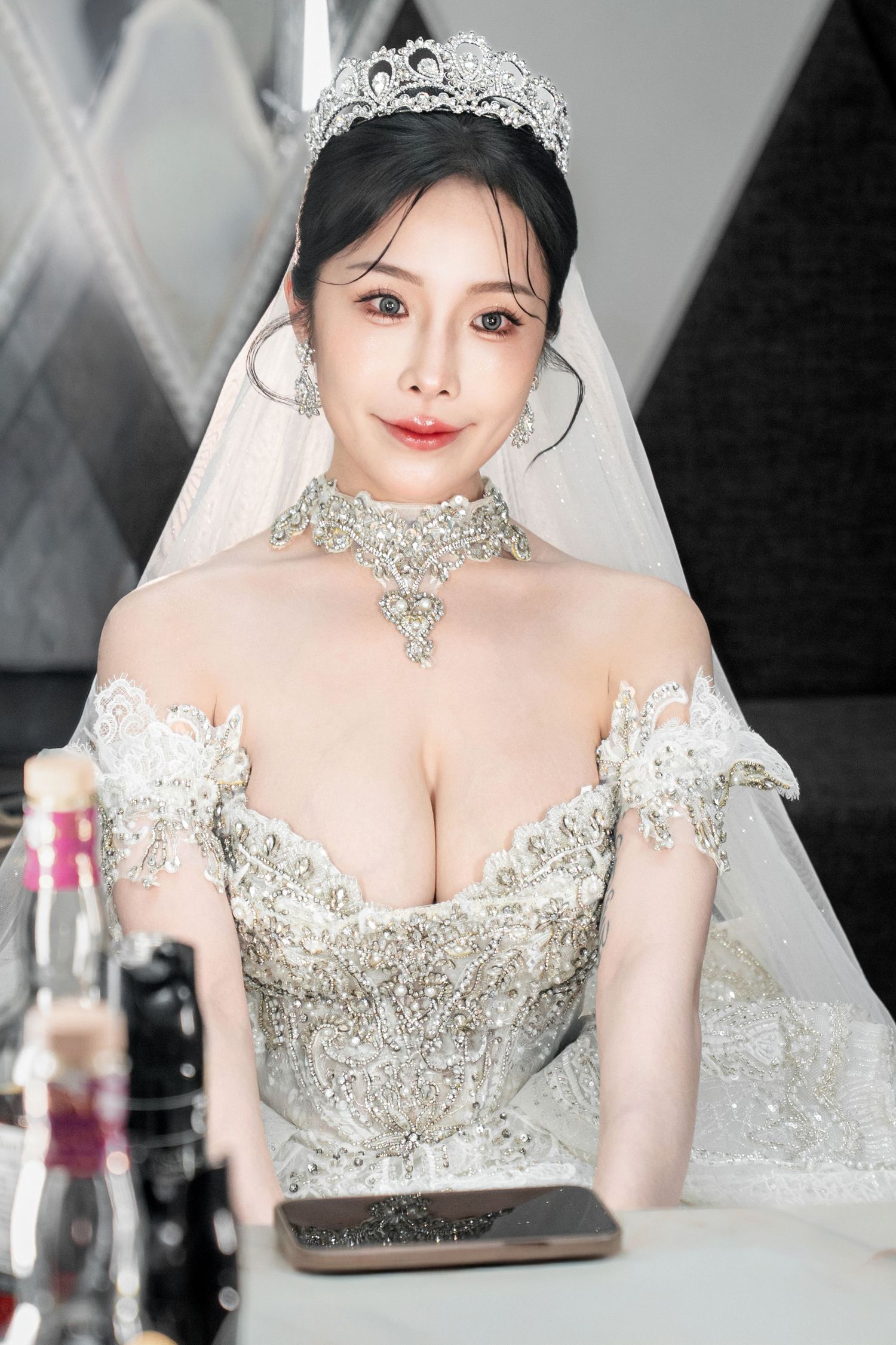 A bride in an ornate white wedding dress with crystal tiara and choker necklace sits at a table.