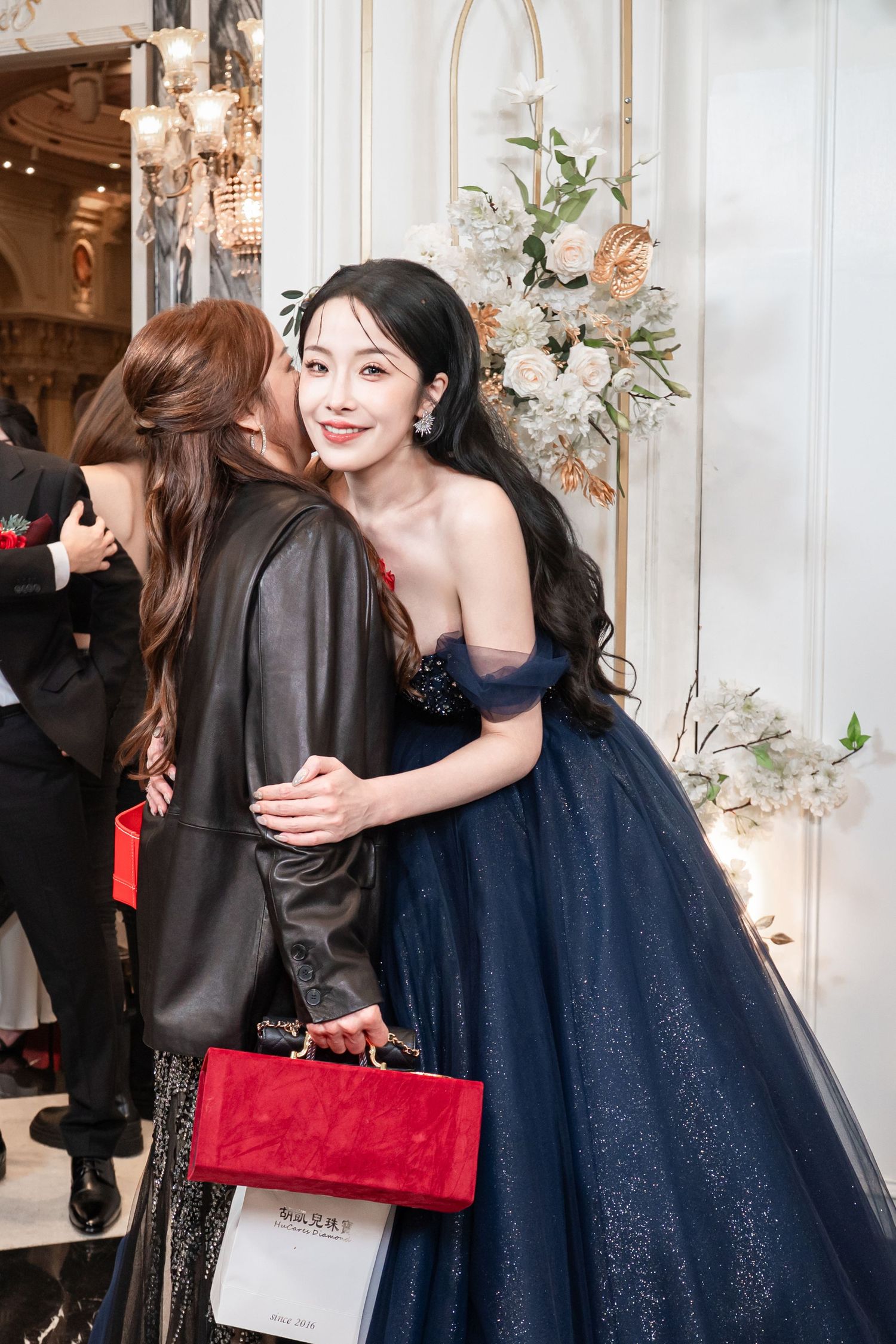 Two people embrace at an elegant event, one wearing a navy blue sparkly ball gown and the other in a black outfit.