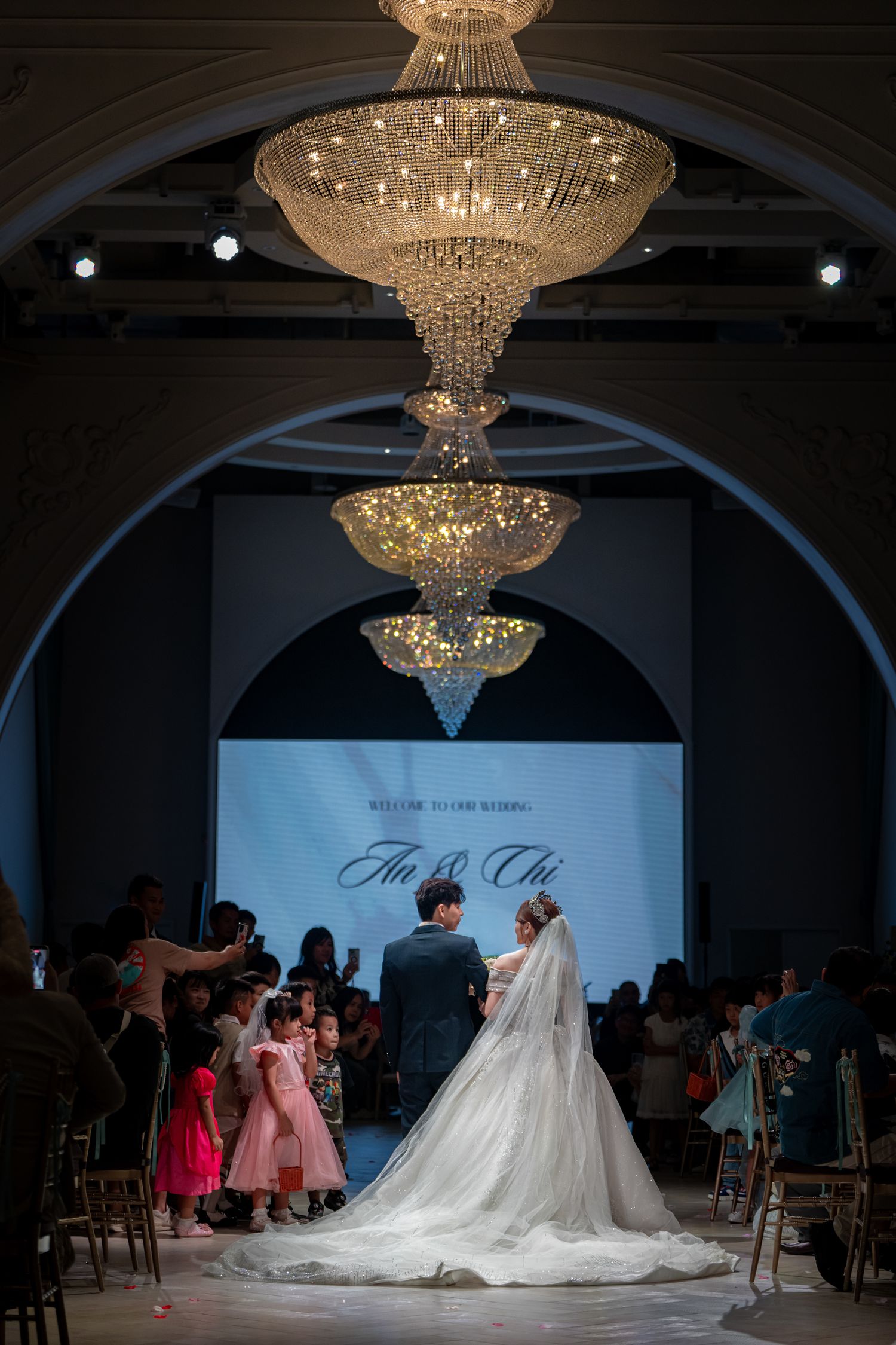 Elegant wedding ceremony under tiered crystal chandeliers, bride in white gown and groom walk the aisle in a grand ballroom.